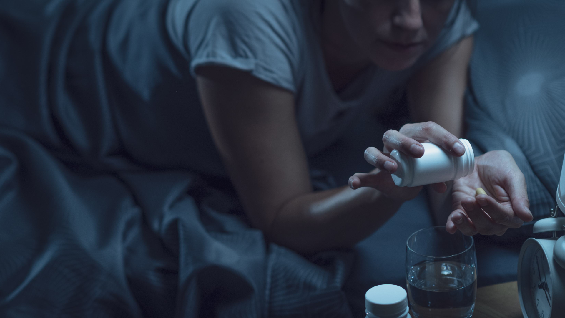 Person pouring melatonin from a bottle into hand next to a glass of water at night.
